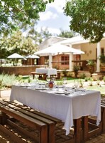Un área de comedor al aire libre con una larga mesa dispuesta para una comida, rodeada de bancos de madera y cubierta por sombrillas.