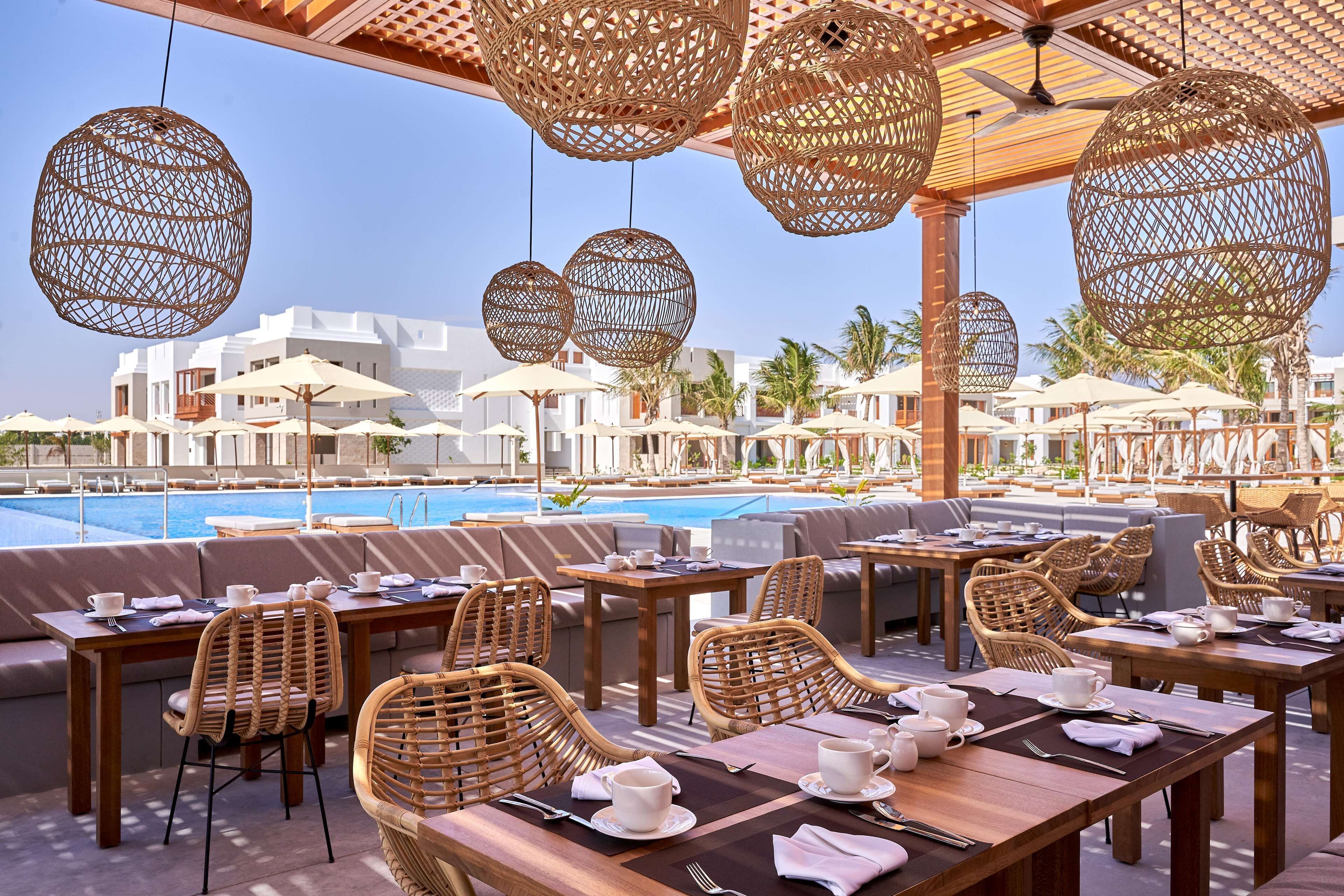An outdoor dining area with wicker chairs and tables, a pool, and white buildings in the background.