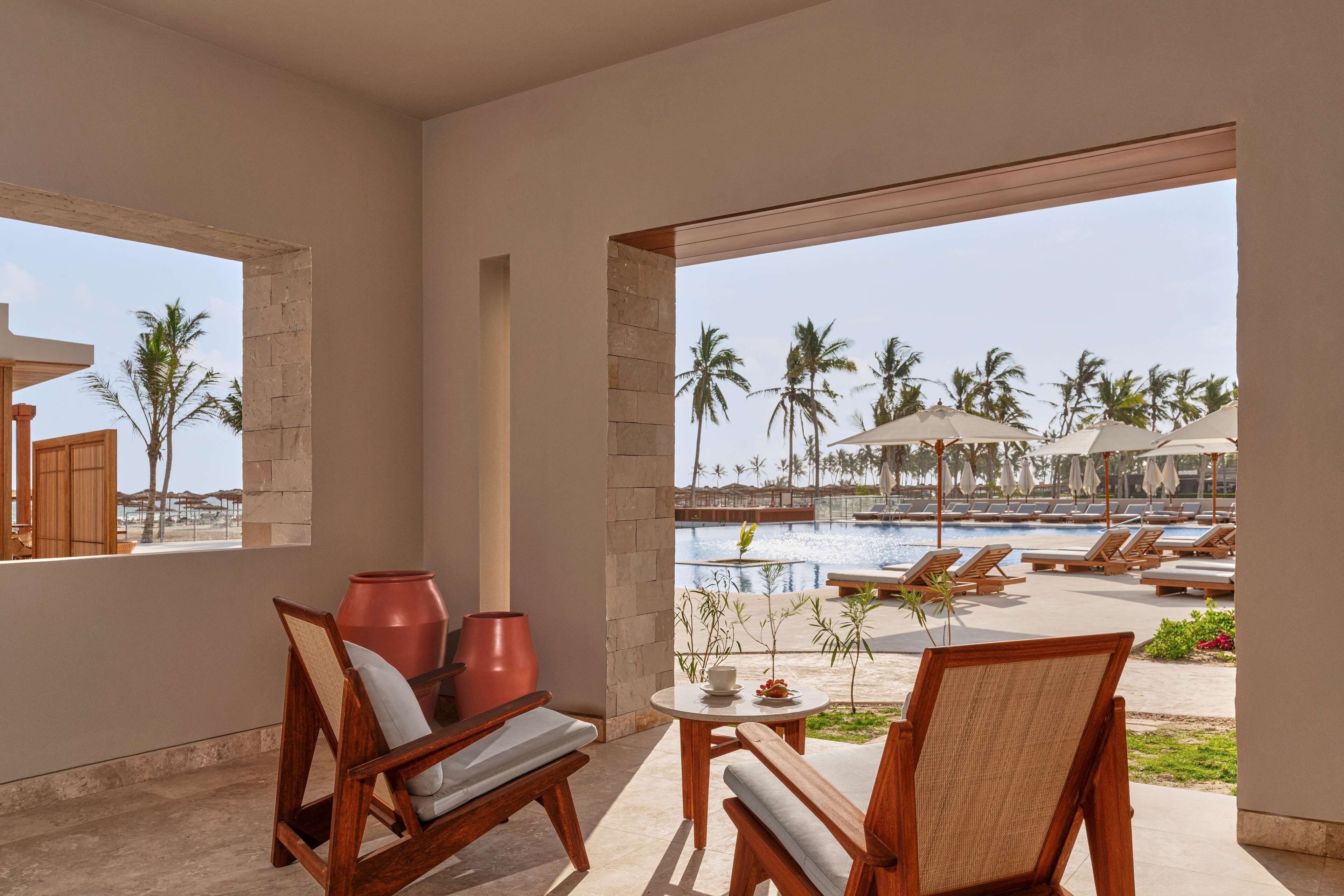 A room with a view of a pool and palm trees, wooden furniture, and a small table with a plant.