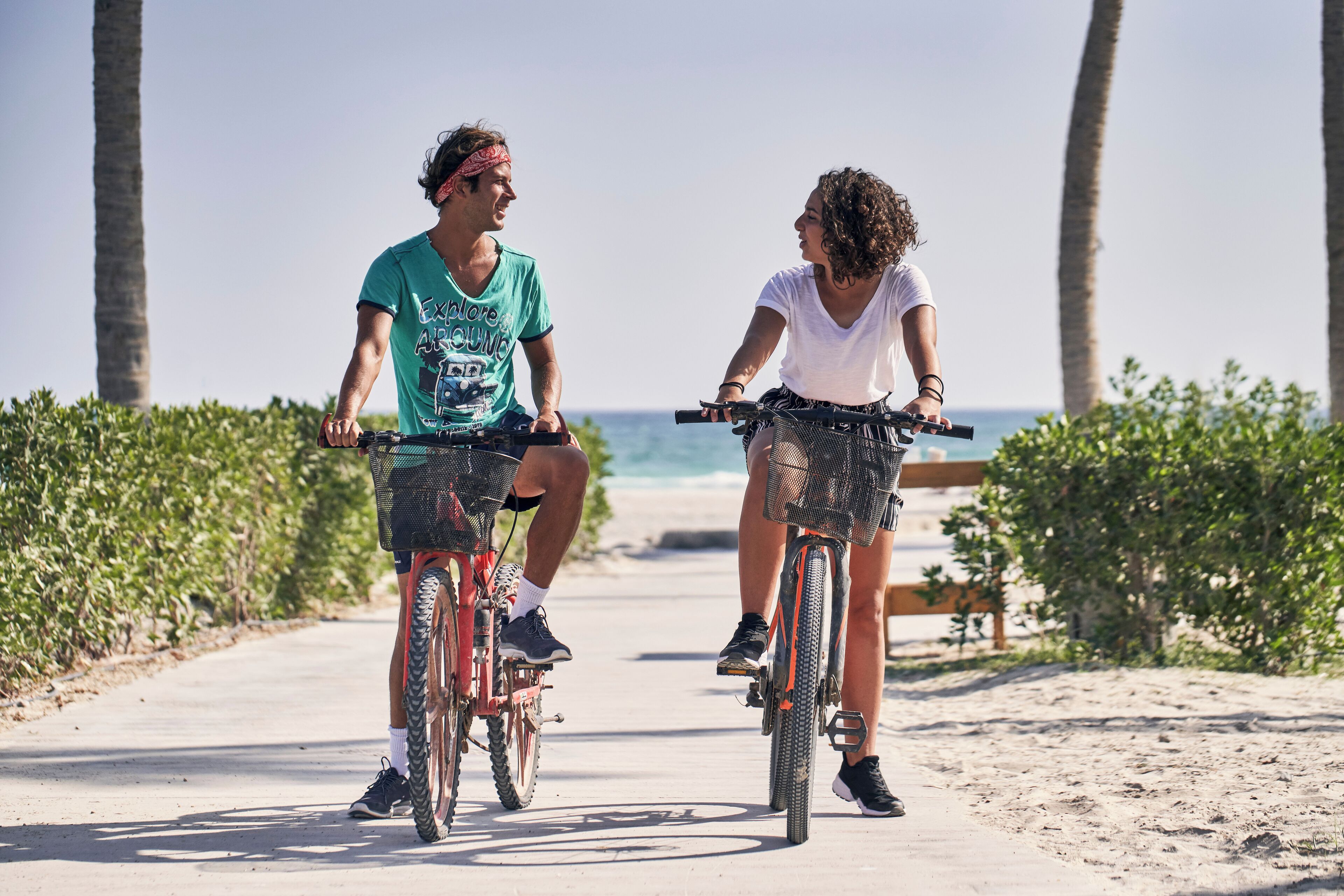 Dos personas andan en bicicleta por un camino pavimentado, con palmeras y un banco al fondo.