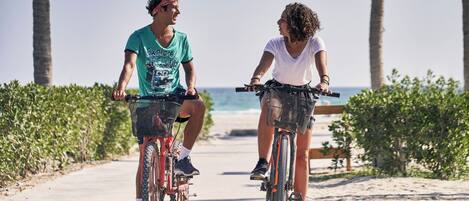 Dos personas andan en bicicleta por un camino pavimentado, con palmeras y un banco al fondo.