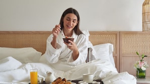 A person in a white robe sitting on a bed with a tray of breakfast items, including toast, a cup, and a glass of orange juice. - The Club by Fanar (Salalah)