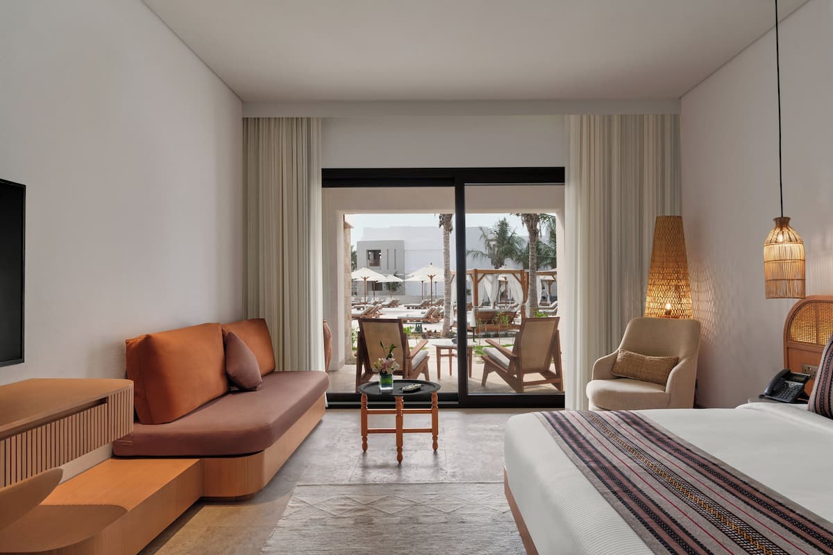 A modern hotel room with a sofa, a chair, a small table, and a bed. There is a view of a pool and palm trees outside.