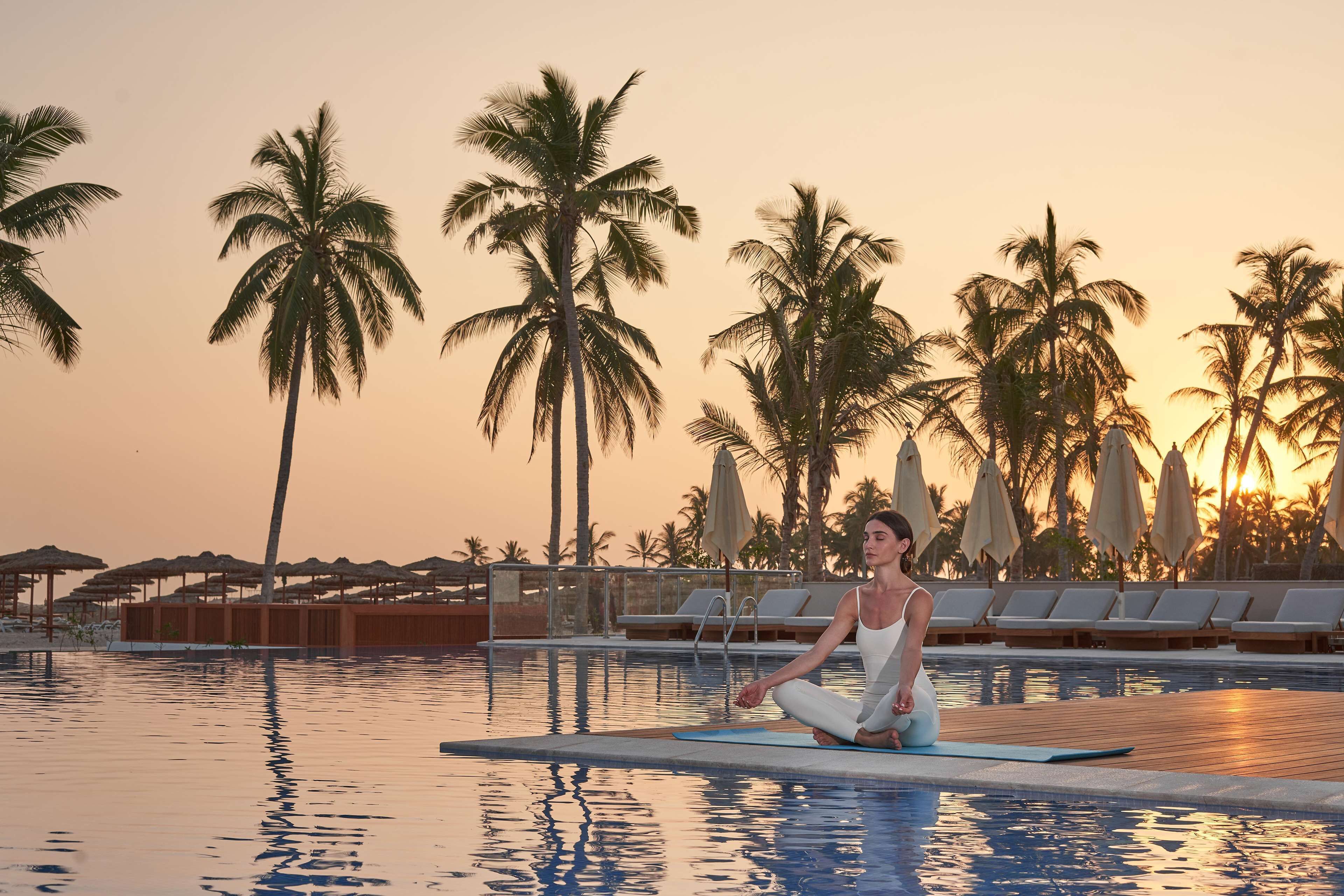 A person meditating by a pool with palm trees and lounge chairs in the background.