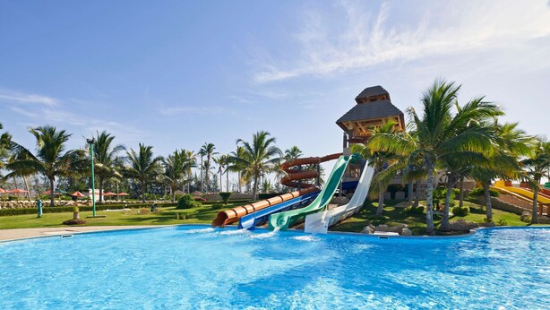 A swimming pool with a water slide, palm trees, and a building with a thatched roof. - The Club by Fanar (Salalah)