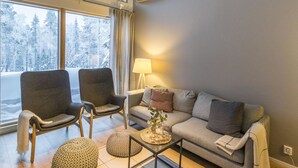 A cozy living room with a sofa, two armchairs, a coffee table, and a lamp. Large windows offer a view of a snowy forest.
