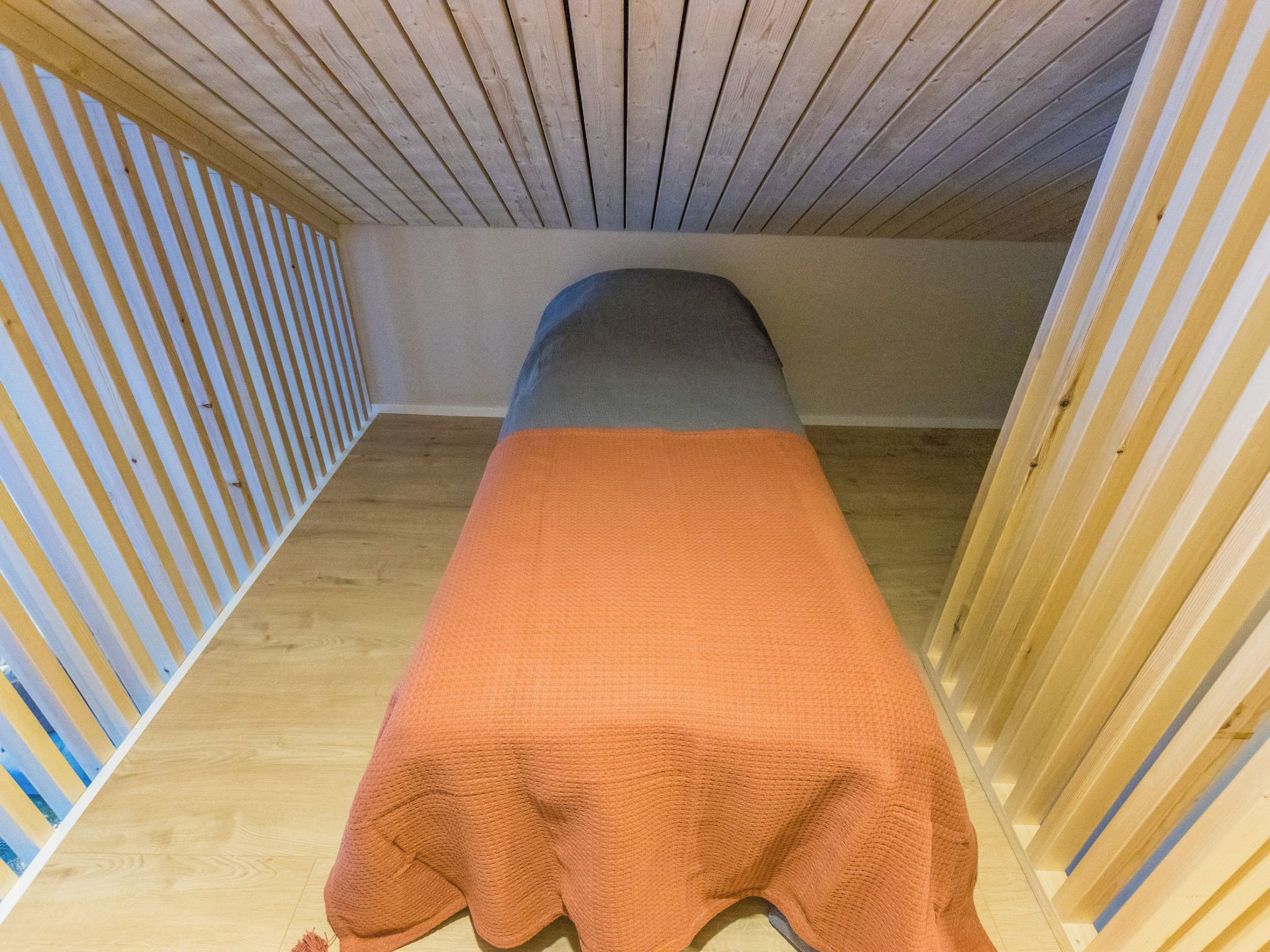 Een eenpersoonsbed met een grijs hoofdeinde in een kamer met houten lambrisering en een houten vloer.