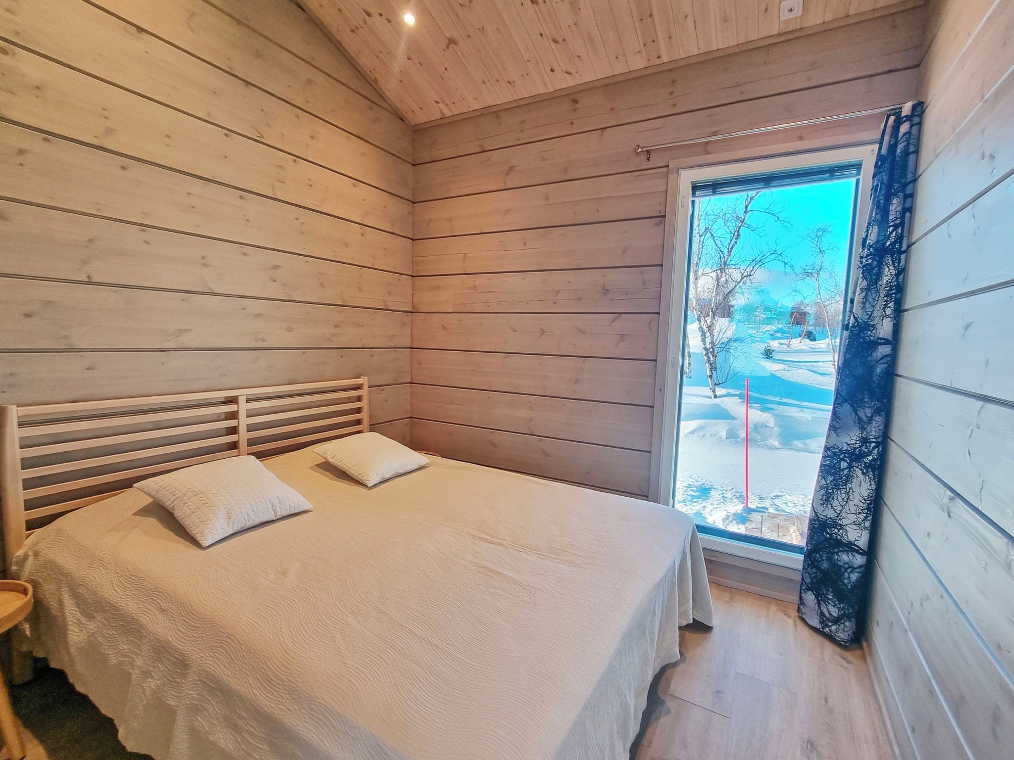 A bedroom with a wooden slatted headboard, two pillows, a bed with a white bedspread, and a window with a view of a snowy landscape.