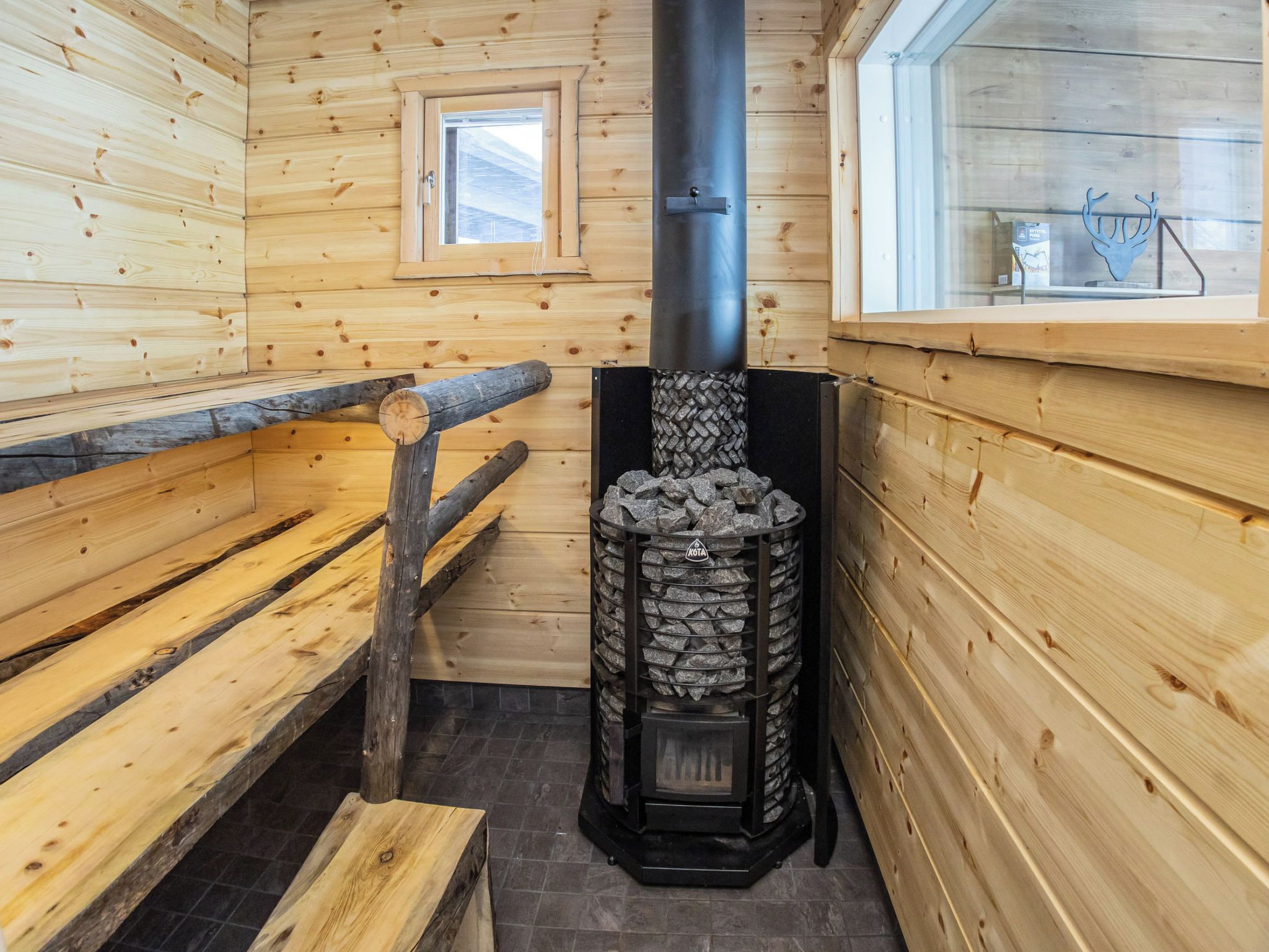 Interior de una cabaña de madera con chimenea de piedra, bancos y una ventana.