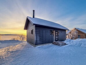 A wooden cabin with a snow-covered roof, a chimney, and a small porch.