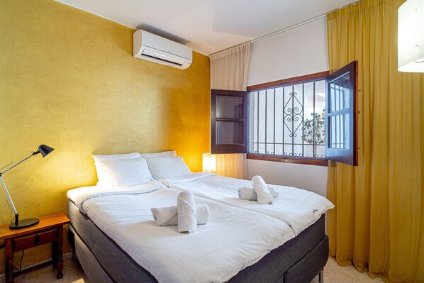 A hotel room with a large bed, a bedside table with a lamp, an air conditioner, and a window with bars.