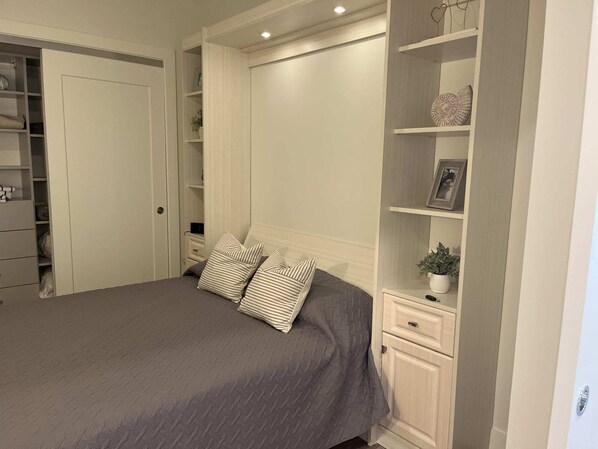 Un dormitorio con cama, almohadas, una mesita de noche, un estante con adornos y un armario.
