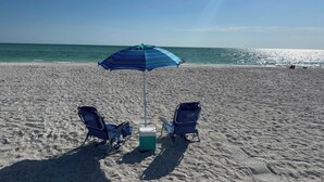 A beach with two blue beach chairs, a cooler, and a striped umbrella. - Luxury 2 Bed 2 Bath Waterfront Condo with Patio Sunsets Gym and Parking (Sarasota)