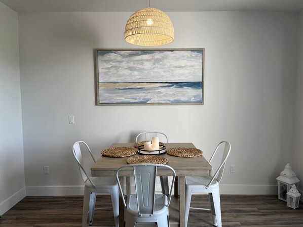 Un comedor con una mesa de madera, sillas de metal, una lámpara colgante de fibras, un cuadro enmarcado y dos faroles blancos.