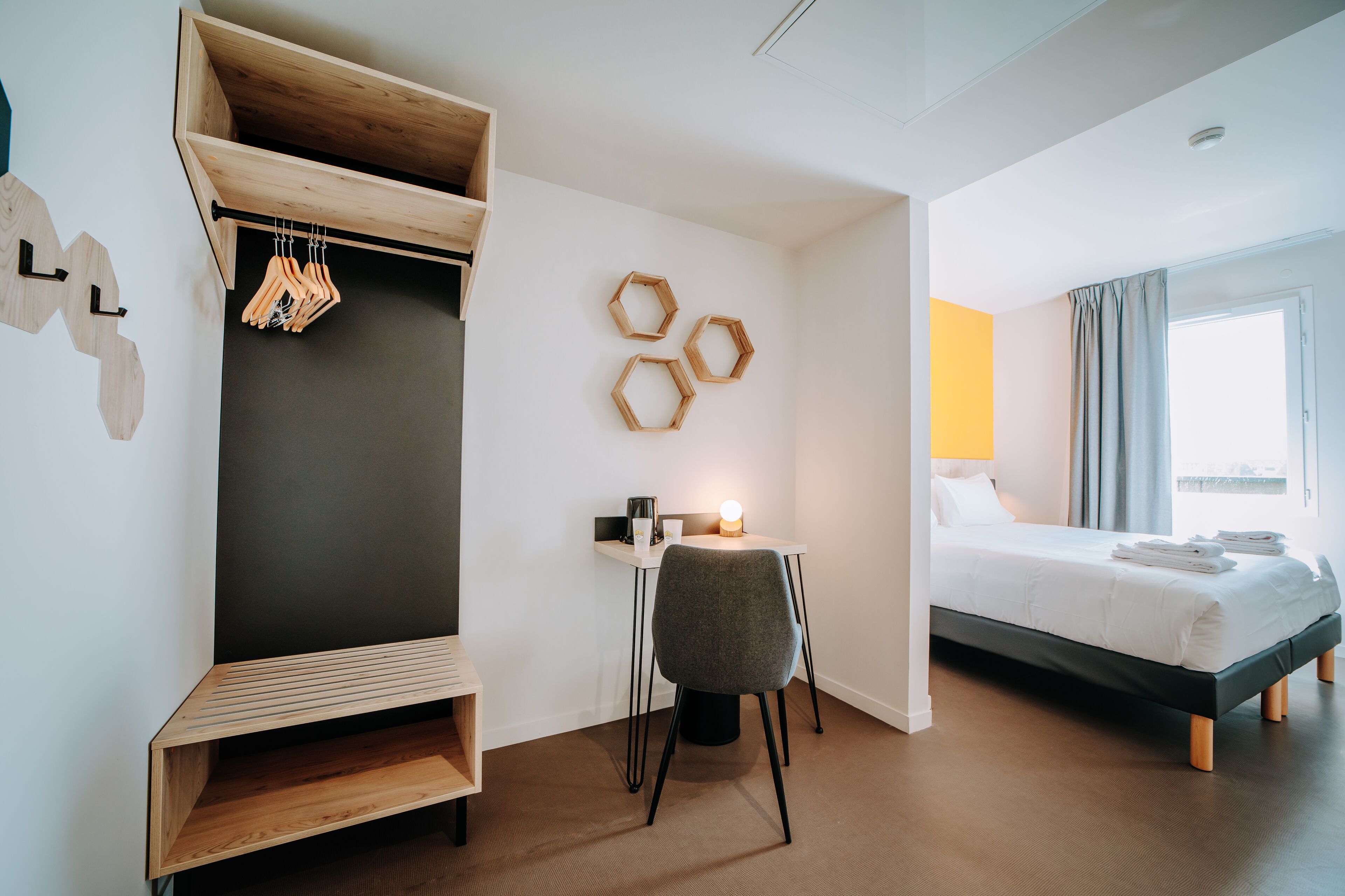 A modern hotel room with a bed, a desk with a chair, a wardrobe, and a wall-mounted coat rack.