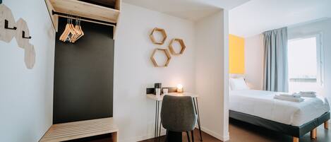 A modern hotel room with a bed, a desk with a chair, a wardrobe, and a wall-mounted coat rack.