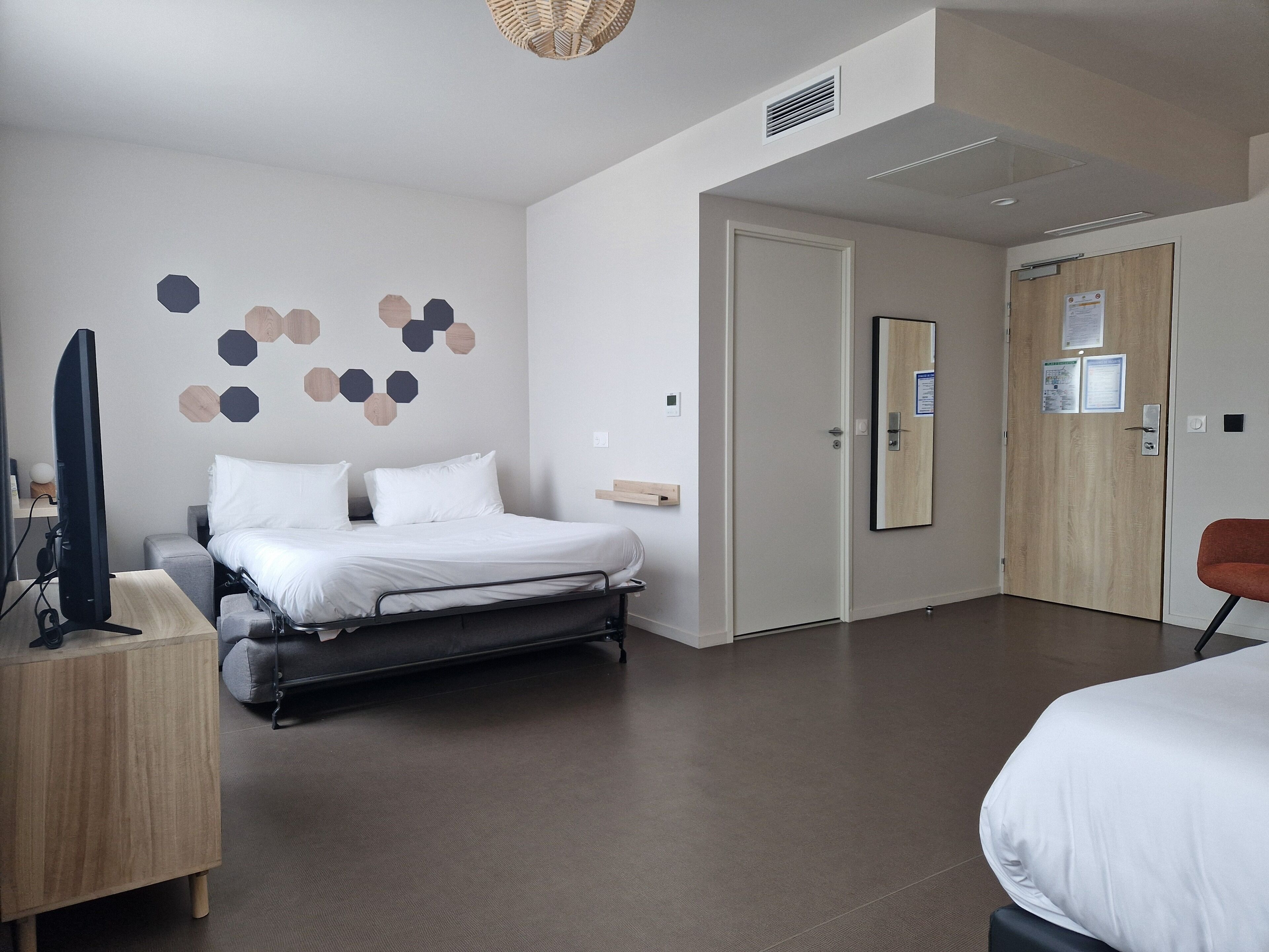 Une chambre d’hôtel moderne équipée d’un lit, d’un bureau avec un ordinateur, d’une télévision, et d’une porte dotée d’un miroir et d’un tableau d’affichage.