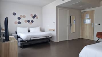 Une chambre d’hôtel moderne équipée d’un lit, d’un bureau avec un ordinateur, d’une télévision, et d’une porte dotée d’un miroir et d’un tableau d’affichage.