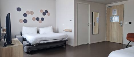 A modern hotel room with a bed, a desk with a computer, a television, and a door with a mirror and a bulletin board.