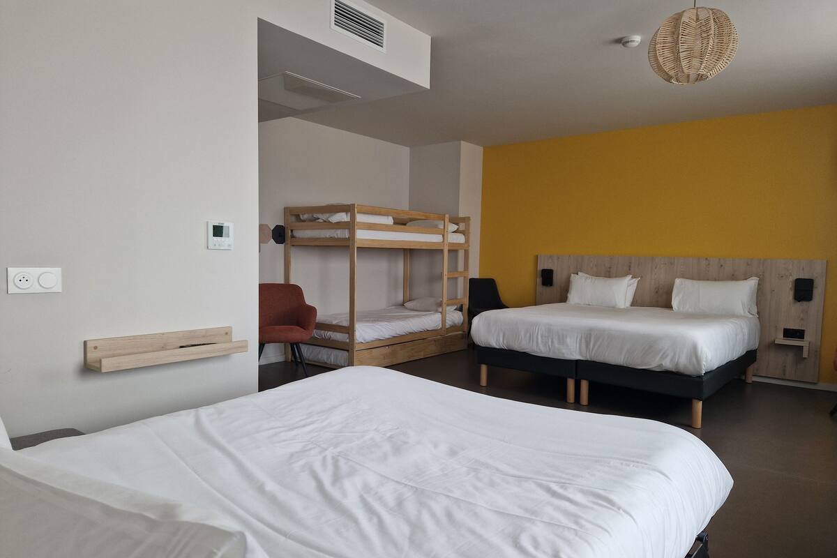 Une chambre d’hôtel moderne avec deux lits, un lit superposé, une chaise en bois, une petite table et un luminaire suspendu.