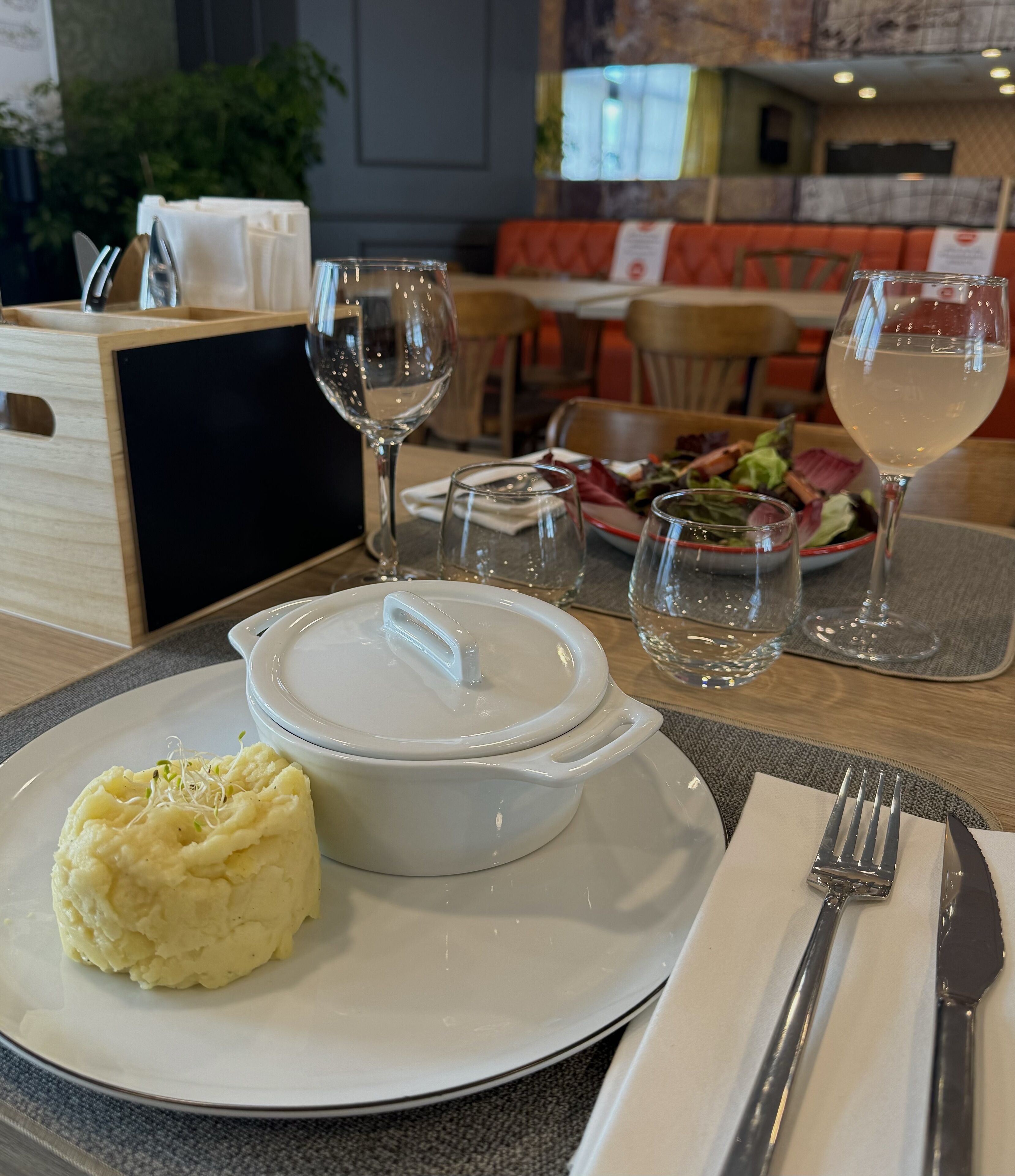 A dining table with a white plate of mashed potatoes and a white bowl, a fork, a knife, and a glass of white wine.