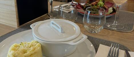 A dining table with a white plate of mashed potatoes and a white bowl, a fork, a knife, and a glass of white wine.