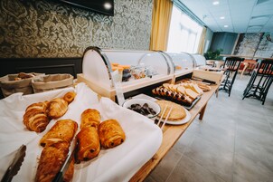 Un buffet garni de viennoiseries, un saladier rempli de fruits et une variété d’aliments disposés sur une table dans une pièce dont les murs sont recouverts d’un papier peint à motifs et qui possède une grande fenêtre.