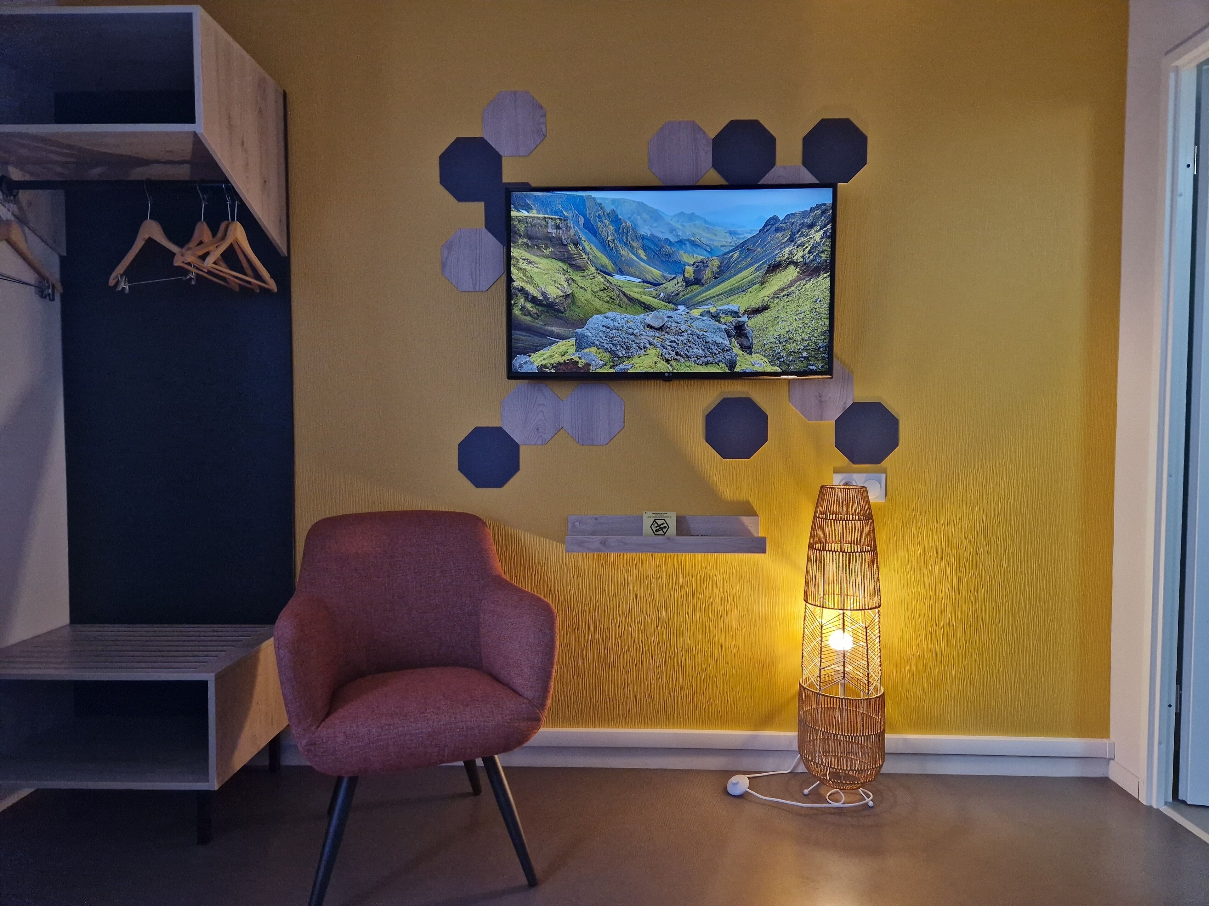 Une chambre moderne équipée d’un téléviseur mural, d’un fauteuil rouge, d’une lampe sur pied et d’une étagère en bois sur laquelle est posé un panneau.