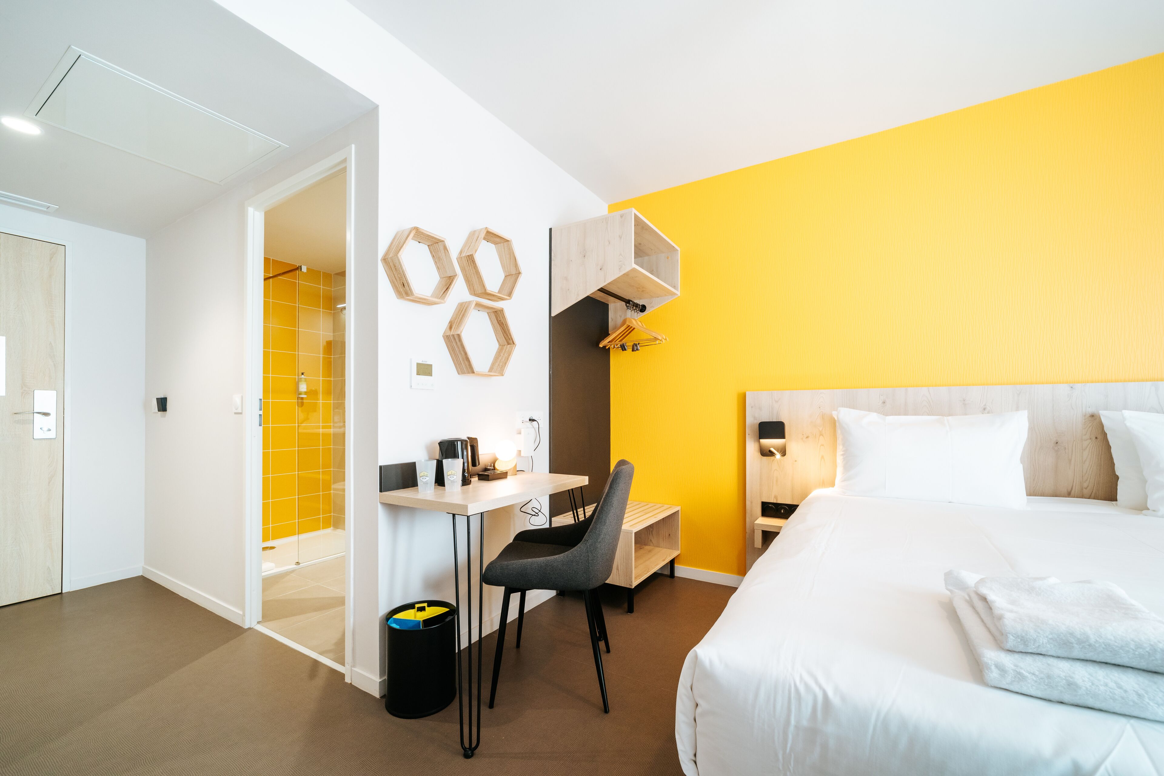 A hotel room with a bed, a desk with a chair, a small table, and a wall-mounted shelf.