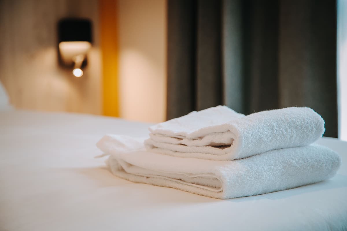 Une pile de serviettes blanches, soigneusement pliées, est posée sur un lit dans une chambre d’hôtel.