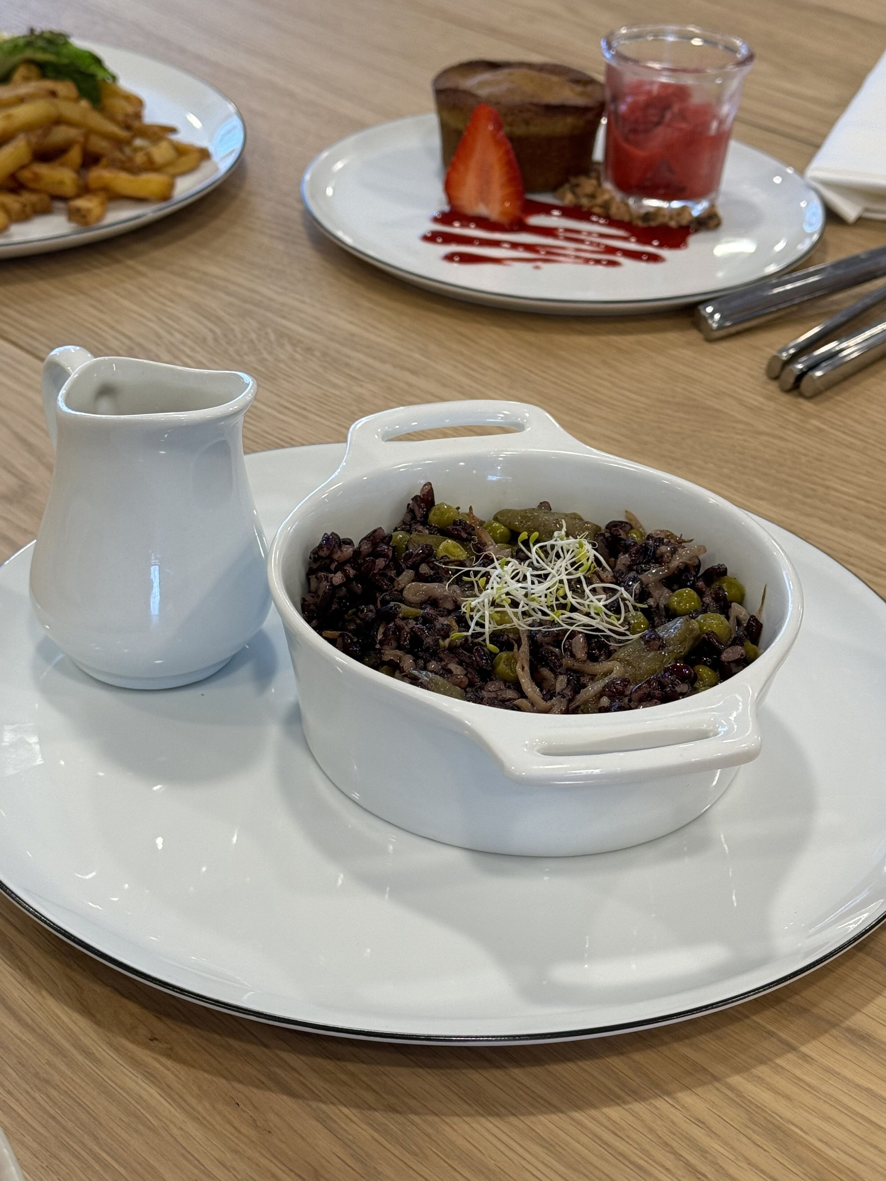 A dining table with a white ceramic bowl containing a dish with black beans and green vegetables, a white pitcher, and plates with food and a glass of red liquid.