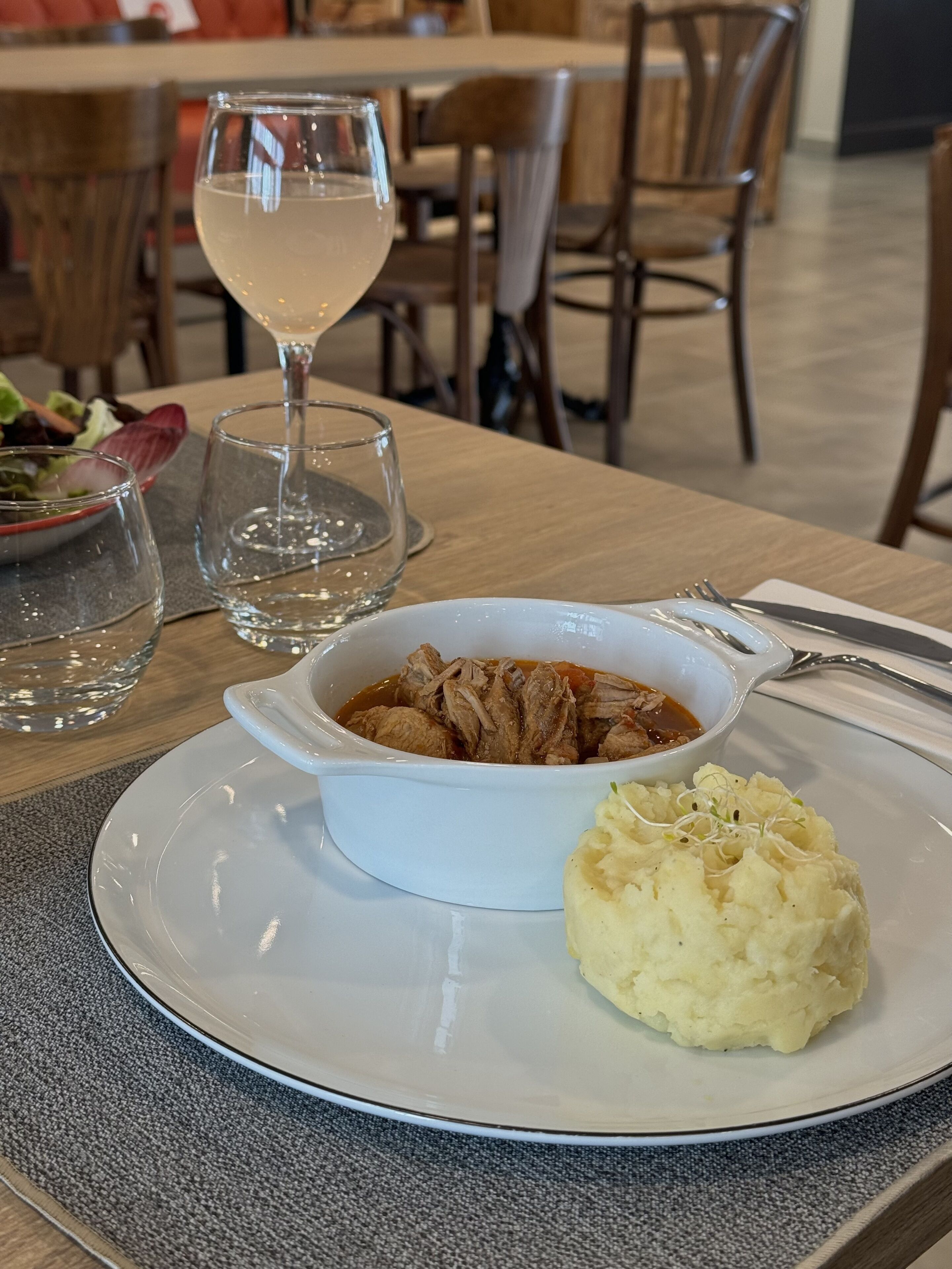 Uma mesa de jantar com um prato branco contendo uma tigela de carne e purê de batata, um copo de vinho branco e uma salada.
