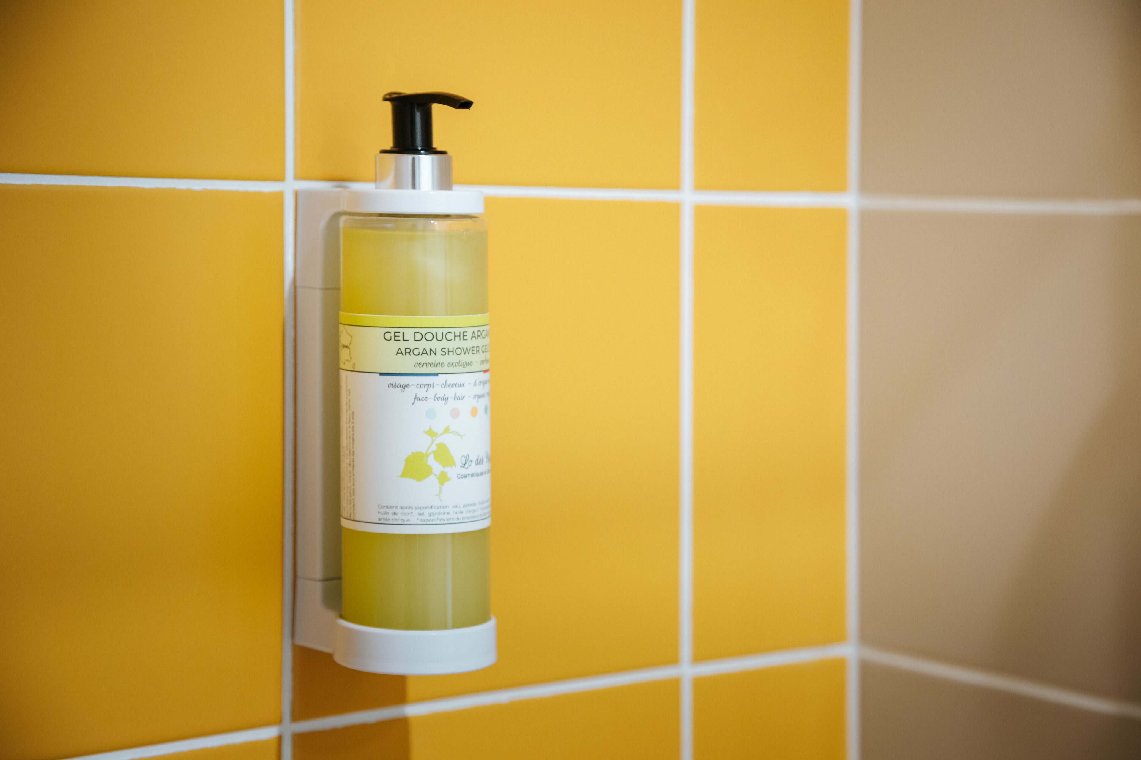 Una botella de gel douche con aceite de argán colgada en una pared de azulejos amarillos.