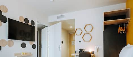 A modern hotel room with a large bed, a flat-screen TV mounted on the wall, and a wooden wardrobe with hanging clothes.