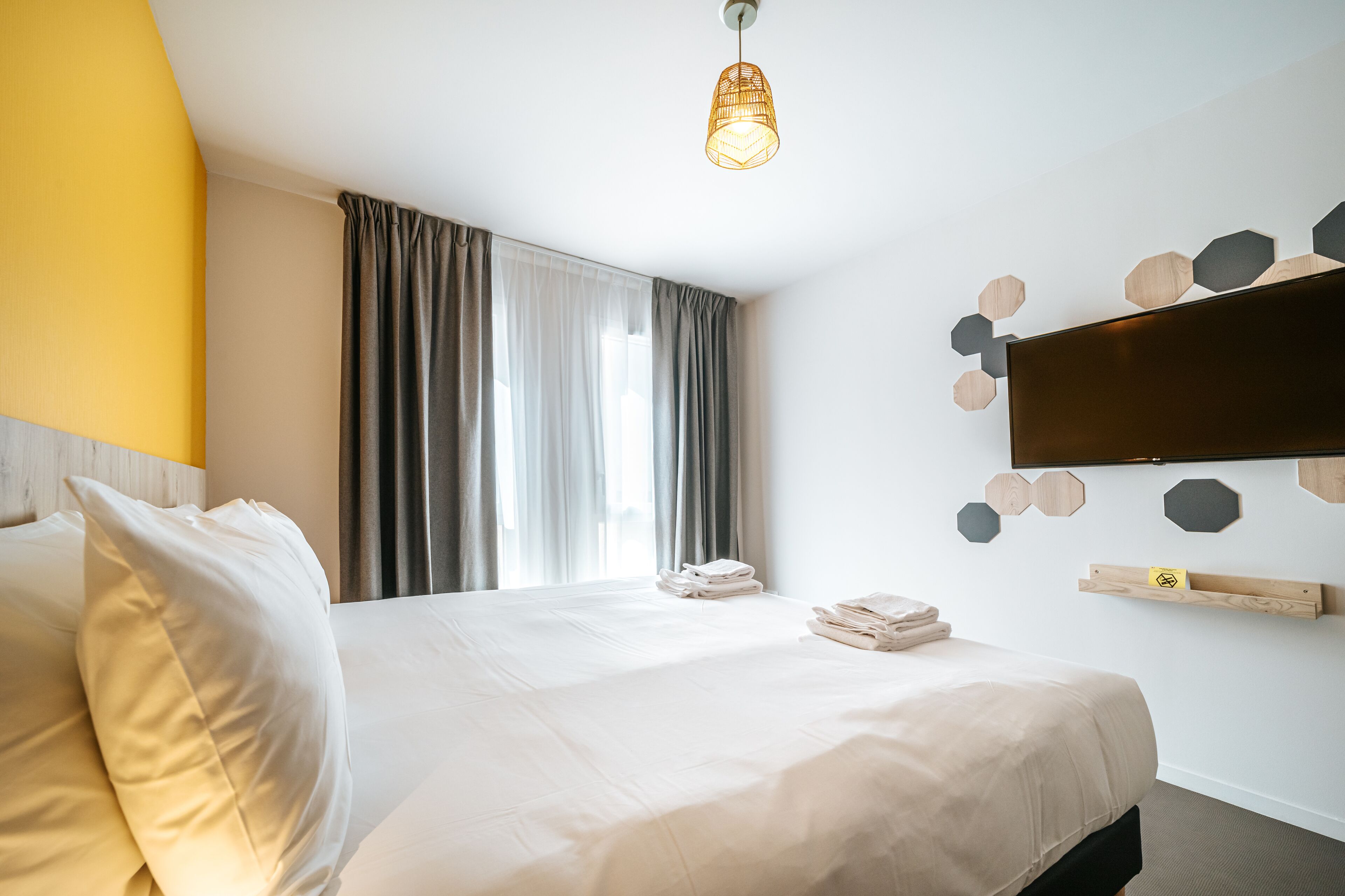 Habitación de hotel con cama, cortinas, televisor montado y estante en la pared.