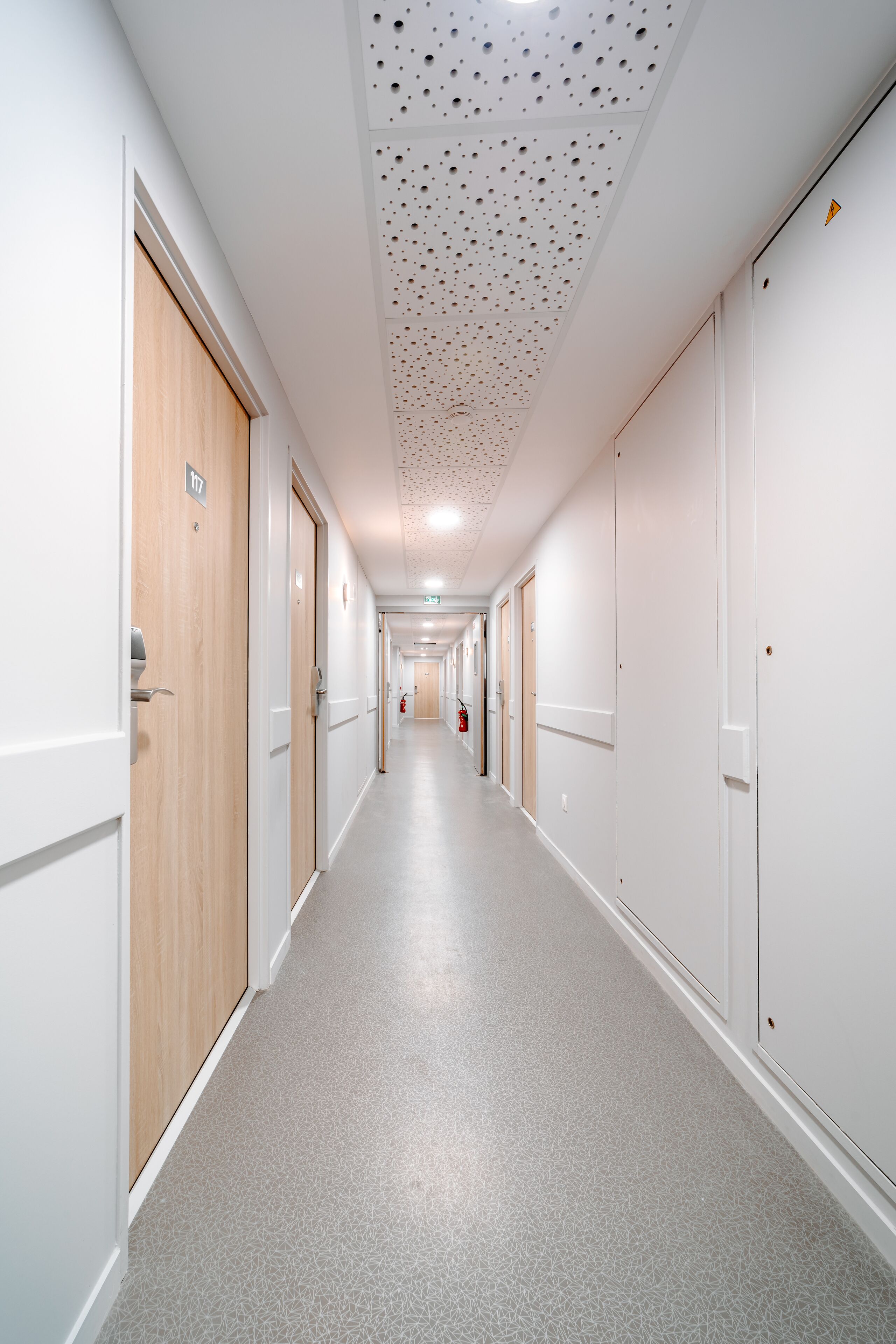 A long, narrow hallway with white walls and doors, a speckled ceiling, and a light-colored floor.