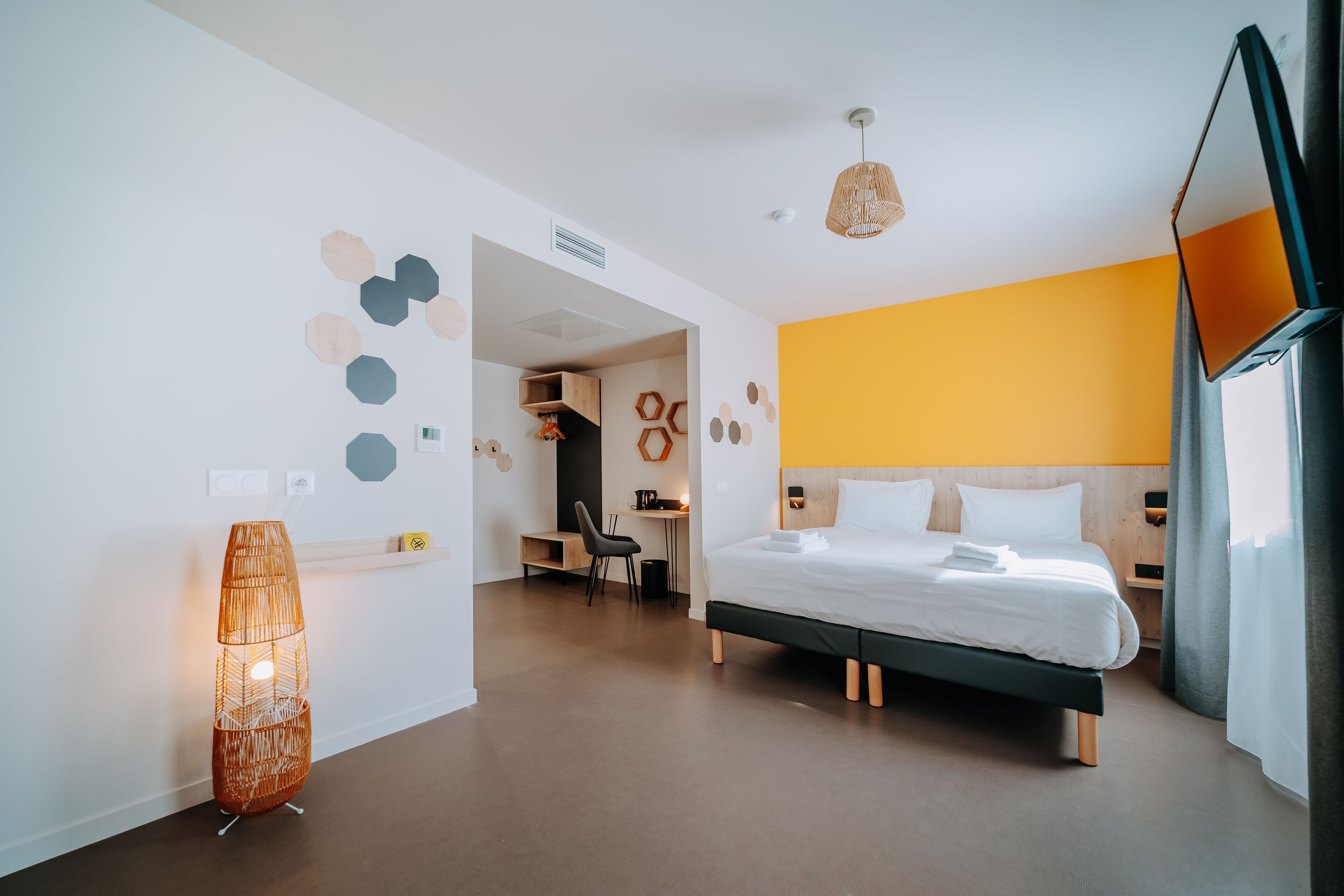 Quarto de hotel moderno com cama, mesa de cabeceira, abajur e vista para o corredor.