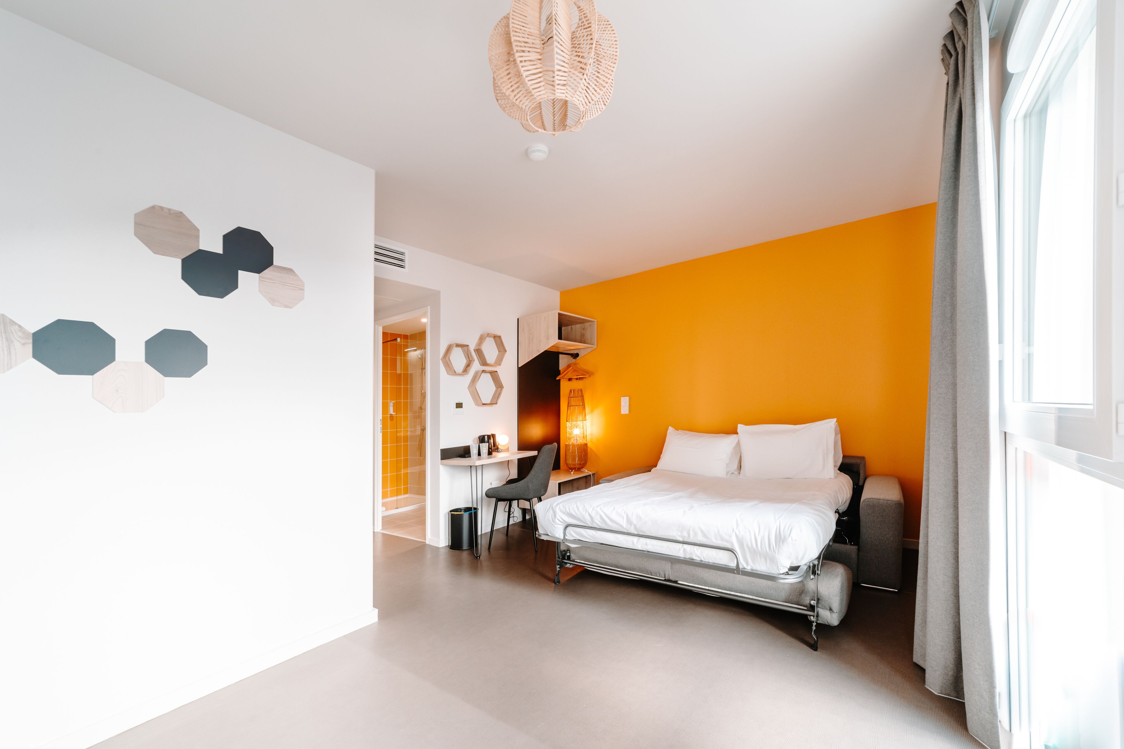 Et moderne hotelværelse med en seng, et skrivebord og en stol. Der er et vindue med gardiner og en væg med hexagonmønstre.
