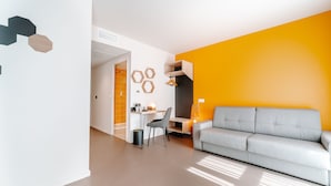 Ein modernes Wohnzimmer mit einem grauen Sofa, einem kleinen Schreibtisch und einem Sessel. Eine Wand ist leuchtend orange und eine andere weiß mit sechseckartigen Verzierungen.