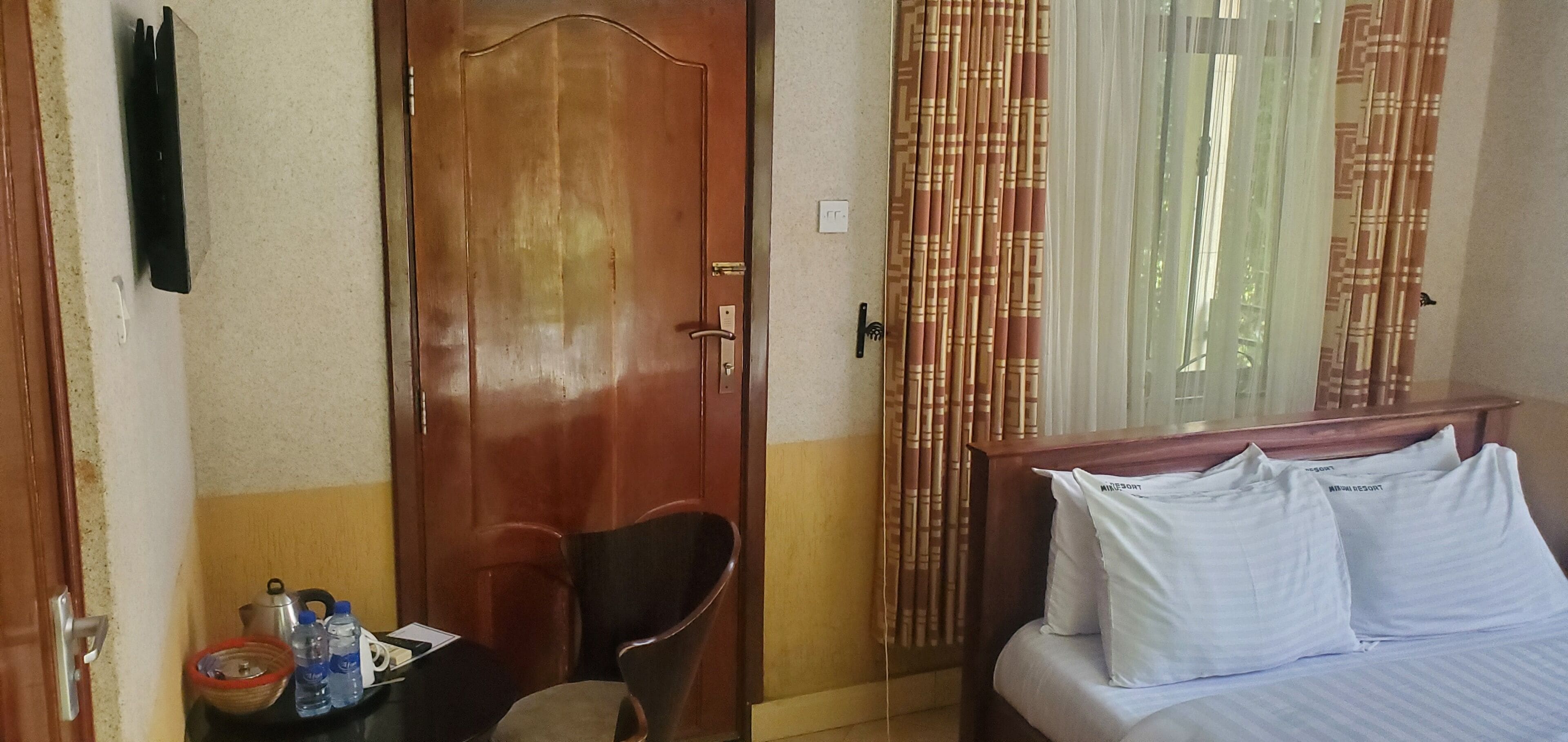 Hotellihuone, jossa on puinen ovi, valkoisilla lakanoiden peitetty sänky, tuoli, pieni pöytä, jossa on hygieniatarvikkeita, ja verhot.