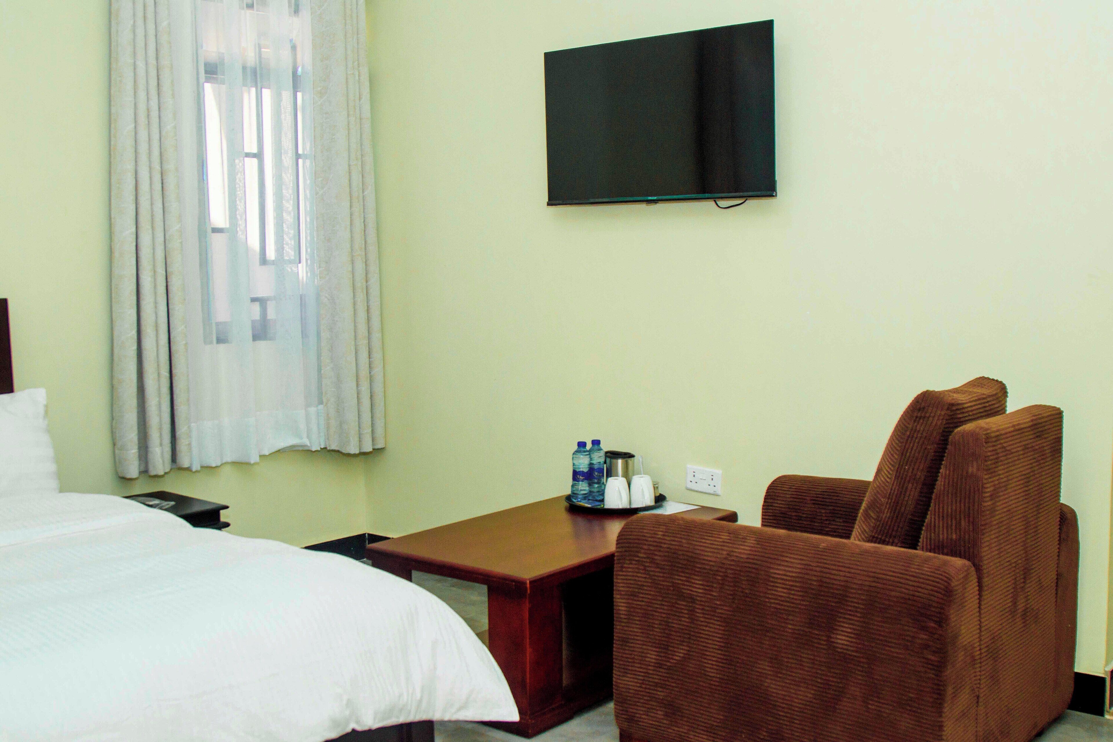 A hotel room with a bed, a brown armchair, a small table with a water bottle and a coffee maker, and a TV mounted on the wall.