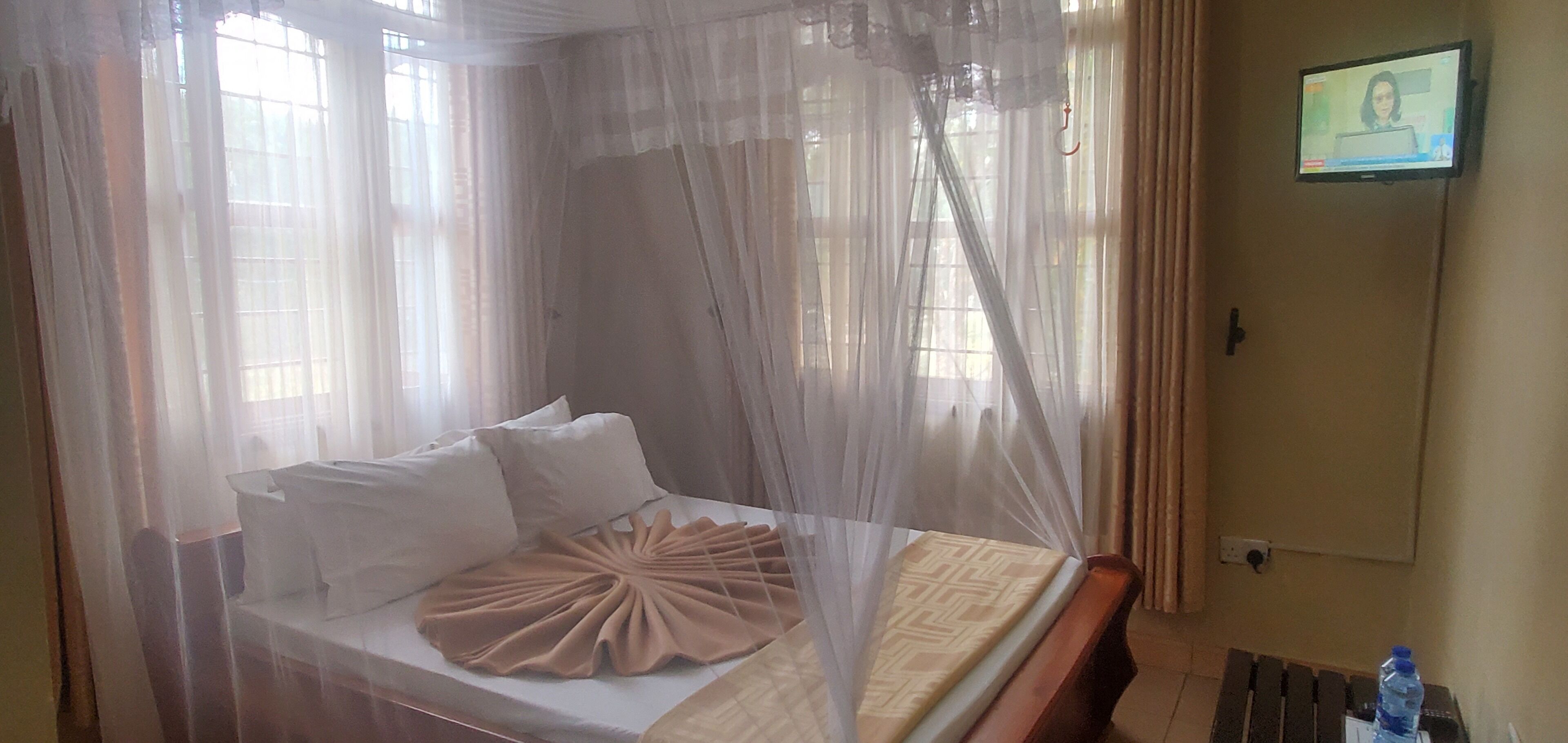 Une chambre d’hôtel avec un lit surmonté d’un dais en voile transparent, un téléviseur fixé au mur et une bouteille d’eau posée sur une petite table.
