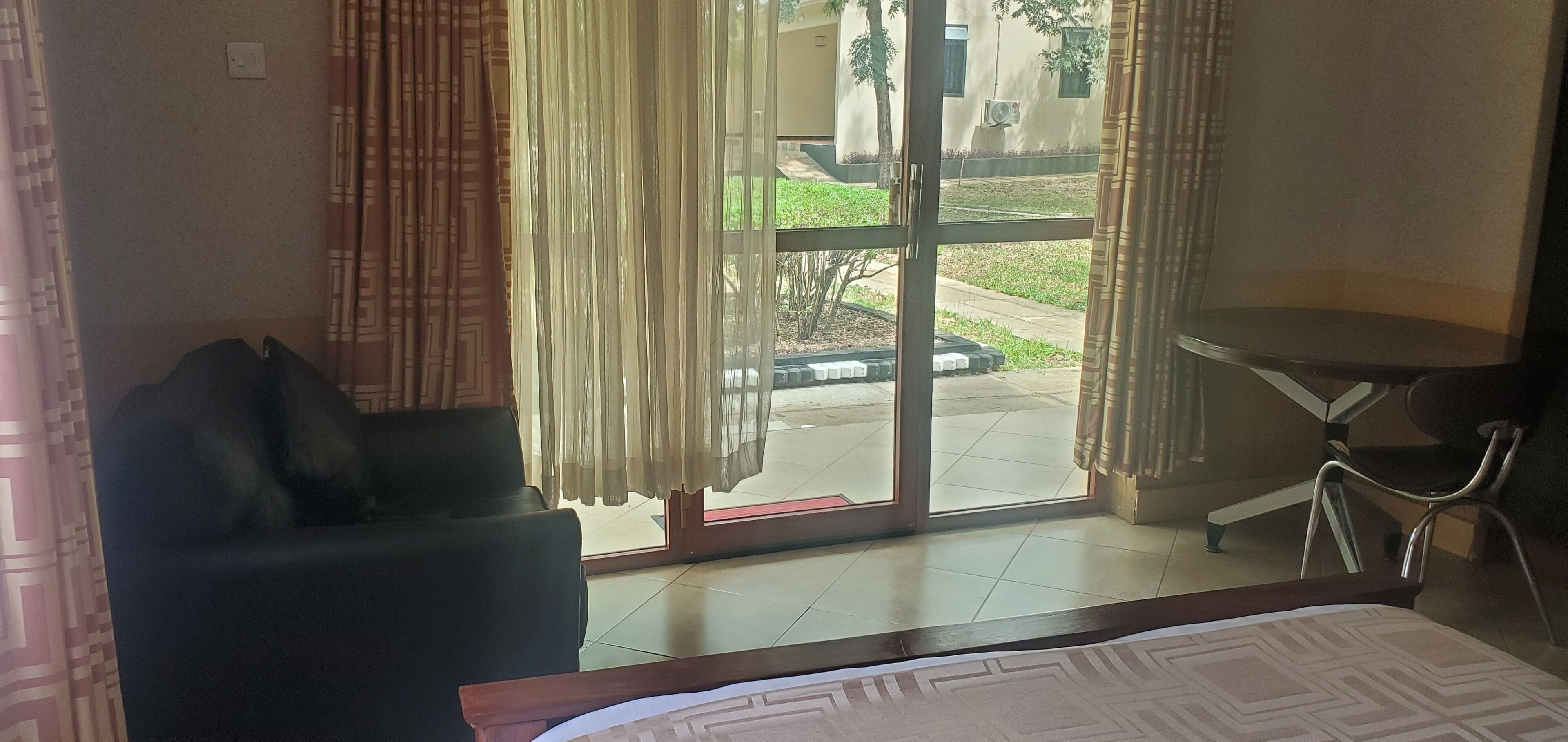 A hotel room with a bed, a black armchair, a small table, and a view of the outdoor area through a sliding glass door.
