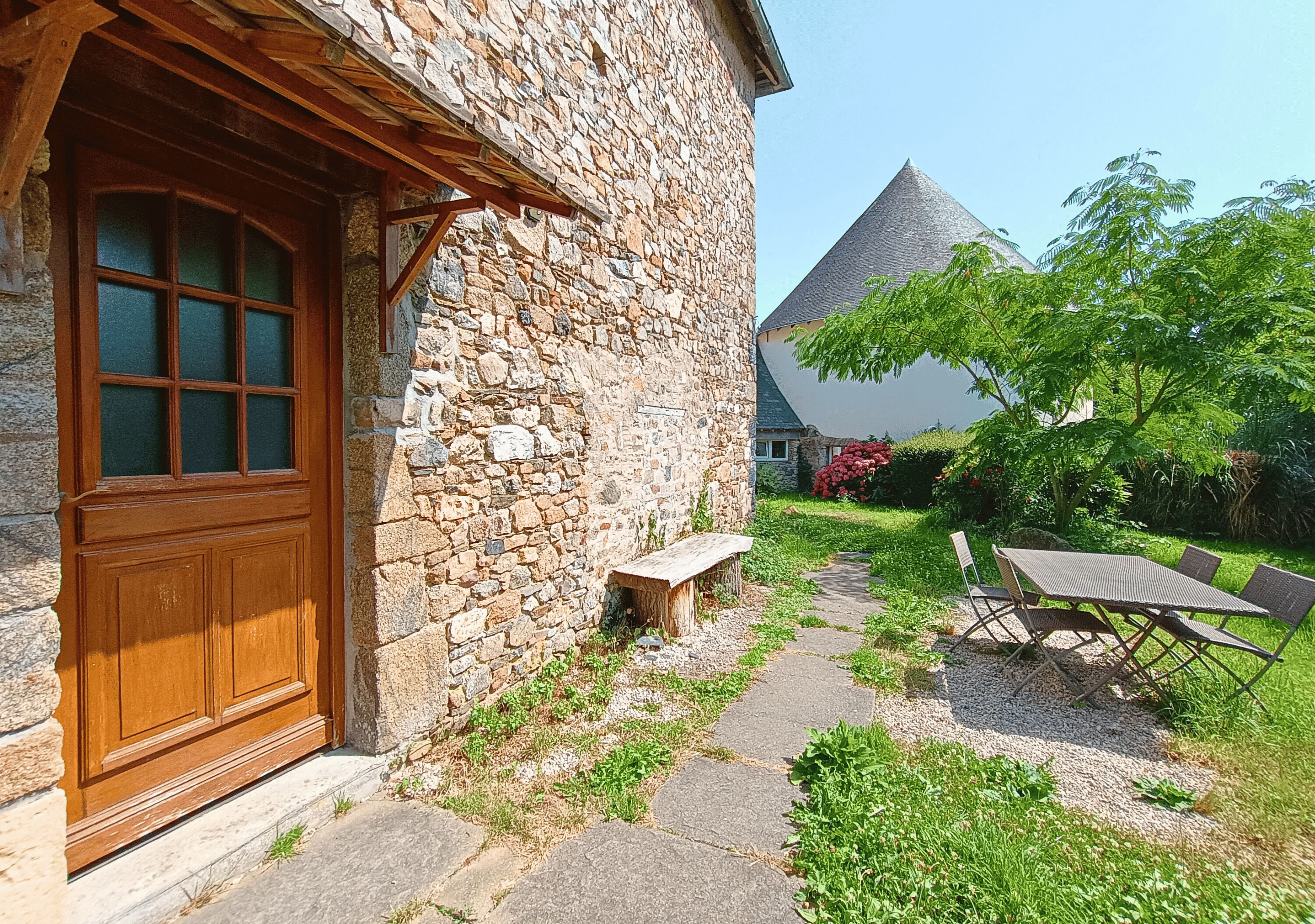 A stone building with a wooden door, a bench, and a table with chairs in a garden.