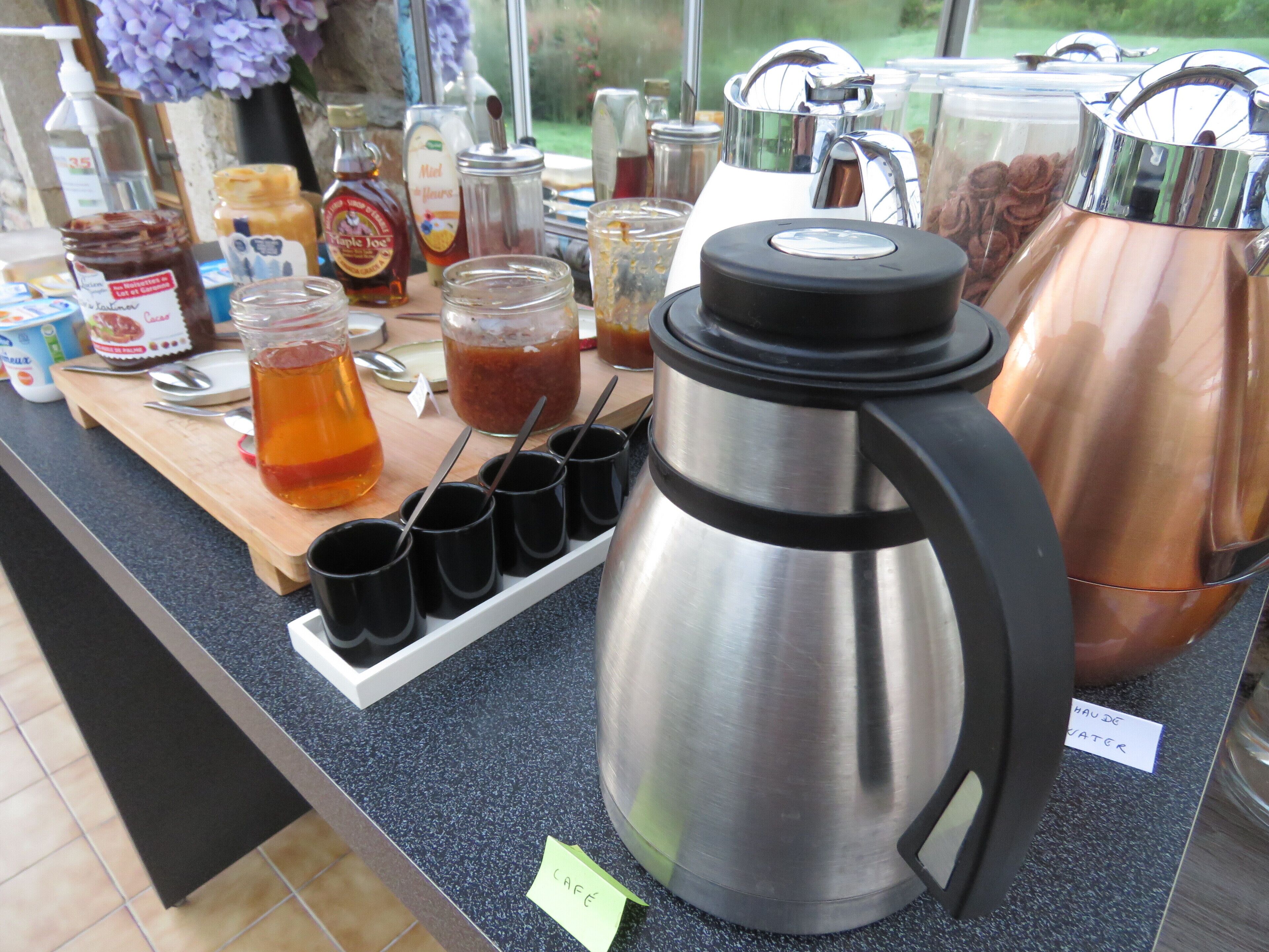 A coffee station with a stainless steel coffee pot, a water dispenser, and various condiments.