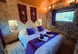 Une pièce avec un mur en pierre, un lit avec une literie blanche et des touches de couleur violette, deux bannières accrochées au mur et une fenêtre donnant sur un paysage.