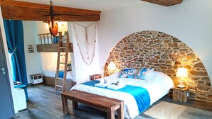 Une chambre avec un mur en arc de pierre, un lit en bois, un banc, une échelle et un filet suspendu.
