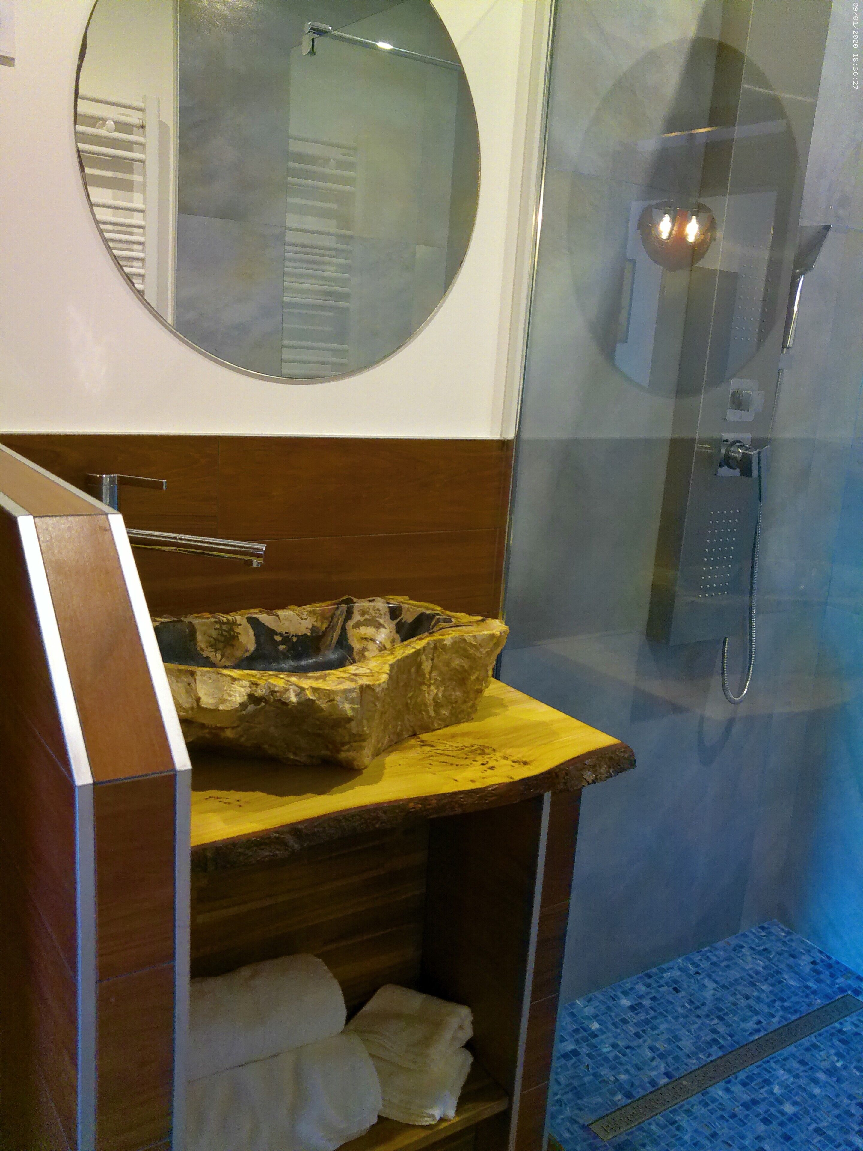 Une salle de bain dotée d’un miroir rond, d’un meuble-lavabo en bois avec un vasque en pierre, d’un porte-serviettes et d’un espace douche avec une porte en verre.