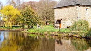 A stone building with a sloped roof near a calm body of water, surrounded by greenery and trees. - La Synchronicité - Chambres d'Hôtes (Saint-Sauveur-Villages)
