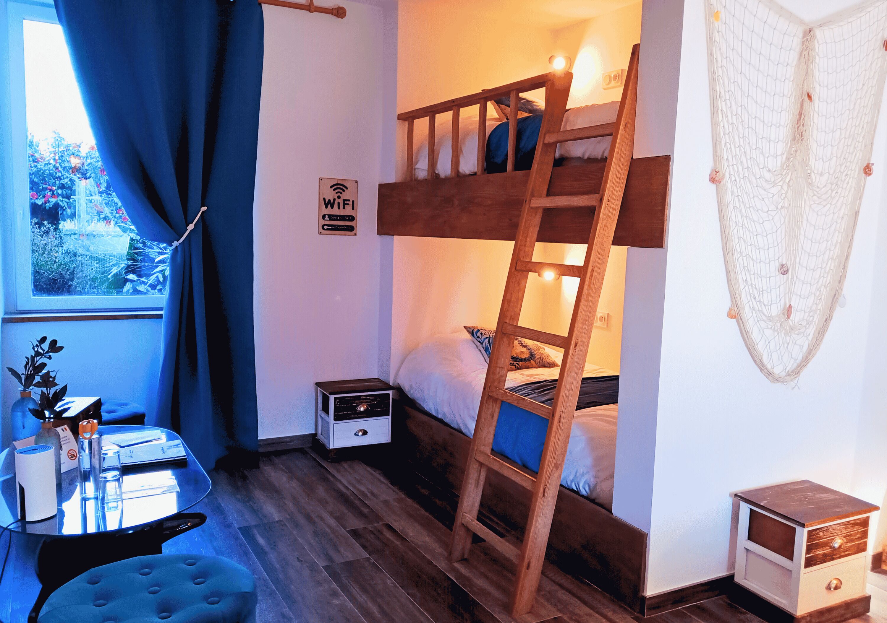 Une chambre avec un lit superposé, une échelle, une petite table de chevet, une prise électrique, un panneau indiquant la présence d’une connexion Wi-Fi et une fenêtre avec des rideaux.